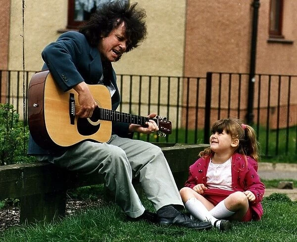 Donovan playing guitar and singing on low wall watched by small girl while on visit to C