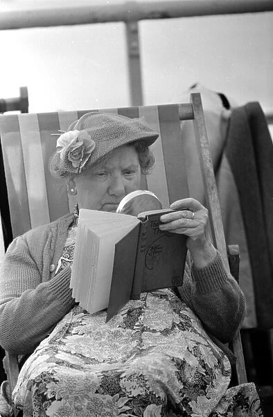 An elderly woman uses a magnifying glass to read her book while sitting on a deckchair