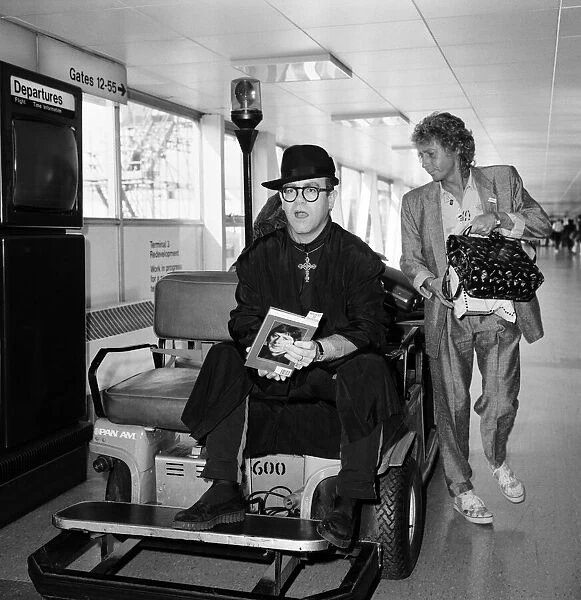 Elton John at Heathrow Airport, wearing a monks black robe and a big gold cross