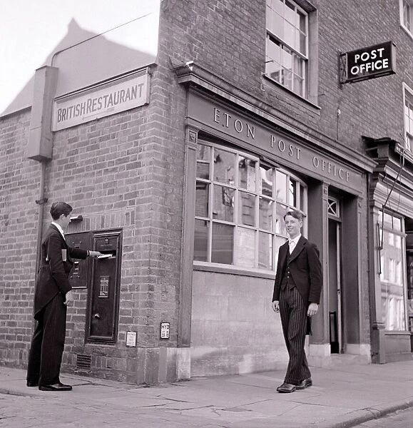 Eton schoolboy walks past Eton Post Office whilst another boy posts a letter dressed in