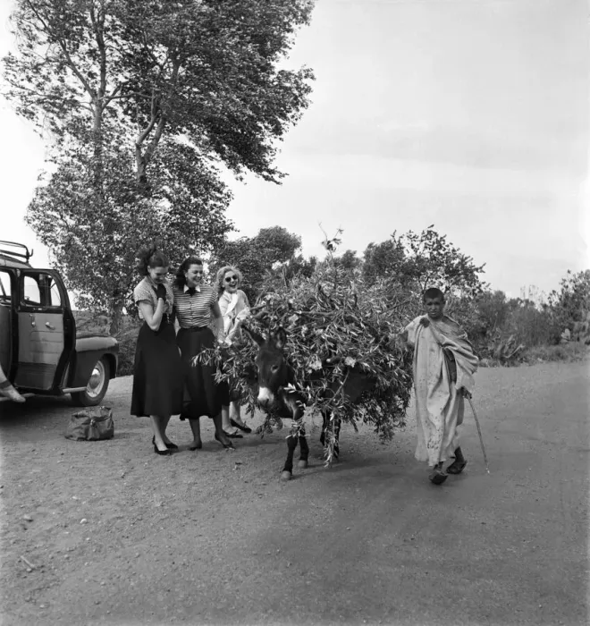 Framed Print : Two French actresses watch as a local man carrying tree branches on his donkey passes by Framed Print : Two French actresses watch as a local man carrying tree branches on his donkey passes by