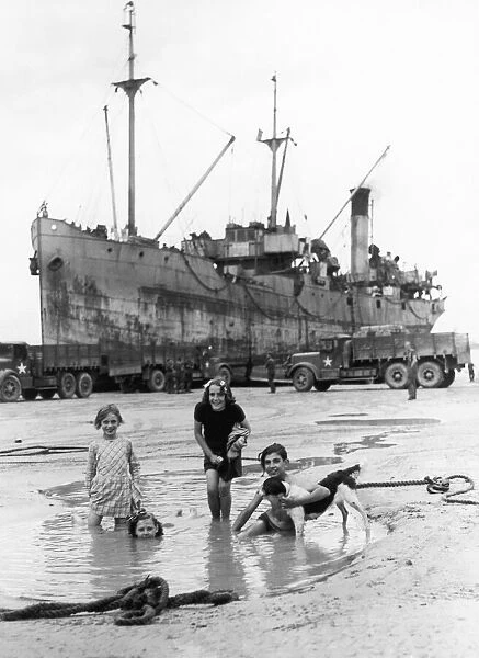 French children playing in a pool on the beach at Arromanches while a ship discharges its