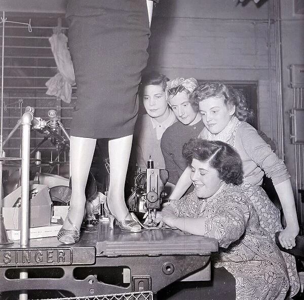 A lady in a pencil skirt stands on a bench while a lady sews on the sewing machine
