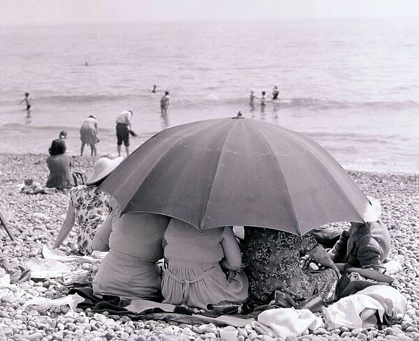 Three large women look out to sea protected under the shade of a large umbrella