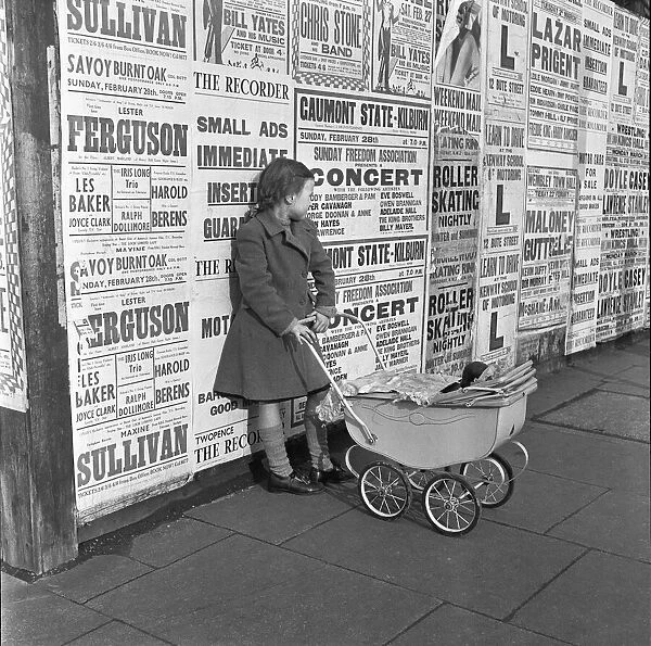 Little girl with toy pram stands waiting for her mother against a background of adverts