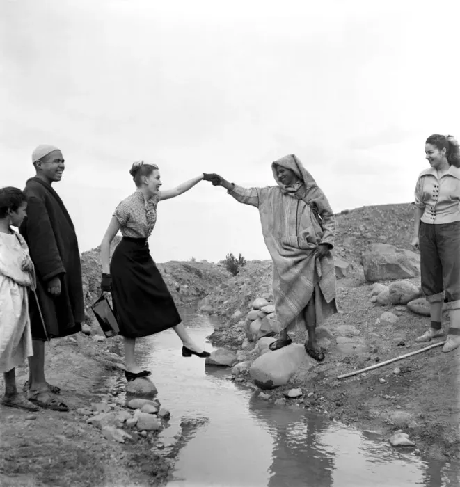 Framed Print : Man in arabic dress lends his hand to help French actresses across a stream as he guides Framed Print : Man in arabic dress lends his hand to help French actresses across a stream as he guides