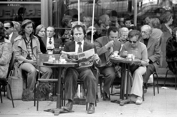 Man reading a newspaper and smoking a cigar as he sits outside in a cafe on the streets