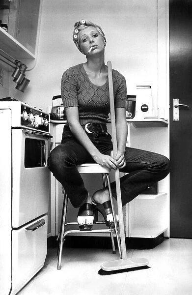 Model Nicki Howarth poses in the kitchen, holding a broom and smoking a cigarette