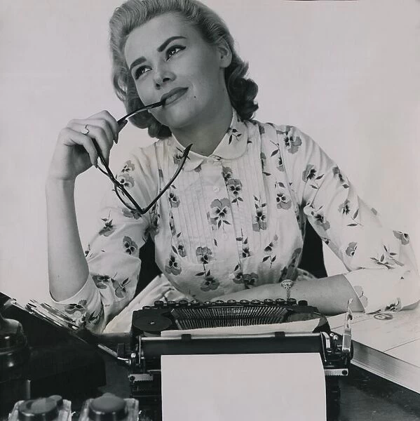 Office worker at typewriter chewing the end of her glasses July 1958