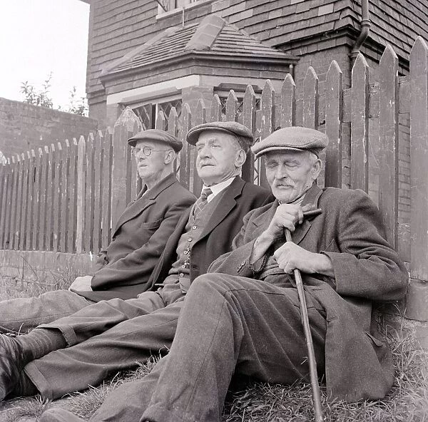 Three old gentlemen rest up against a wooden picket fence and watch the world go by