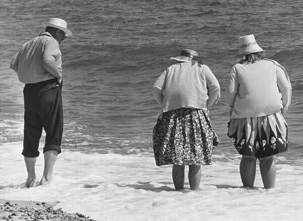 Two old women and one old man with hats stand on the beach with their feet in the water
