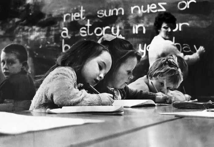 Framed Print : Primary School Children writing at desk with teacher in the background writing on a