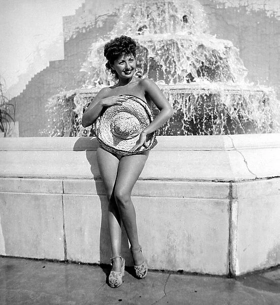 Renee Lester covering herself up with a straw hat as she stands in front of a fountain at