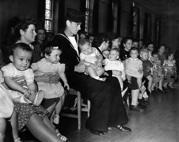 A sailor sits in a hall with his child on his lap with a room full of mothers