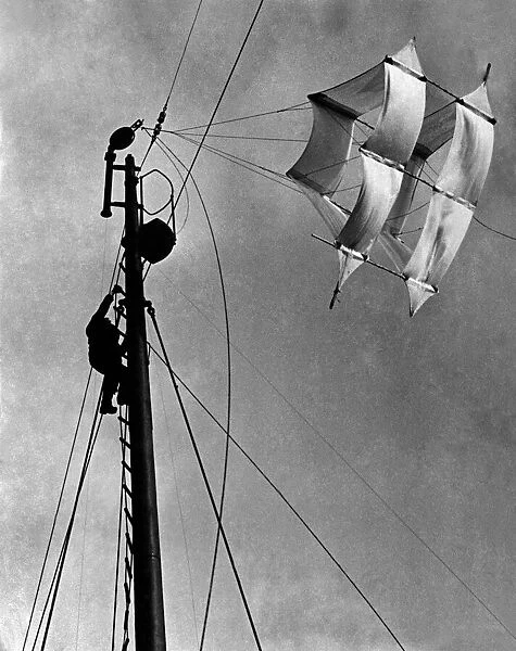 A seaman up the main mast adjusting the huge kite that carries a slender cable