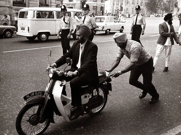 A Sikh man riding a motorcycle, wearing a turban. pushes by a fellow sikh