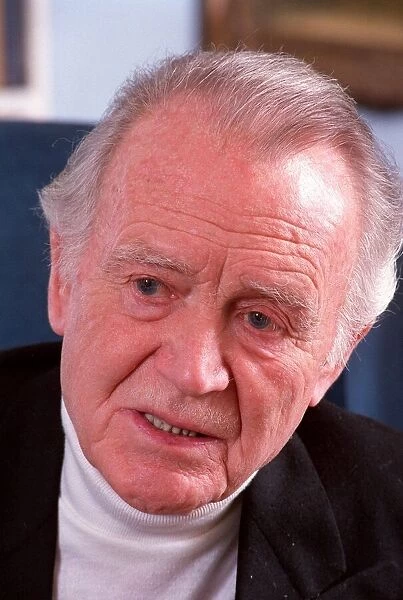 SIR JOHN MILLS IN PHOTO SHOOT AT HIS HOME SITTING IN ARMCHAIR WITH BOOKCASE AS BACKGROUND