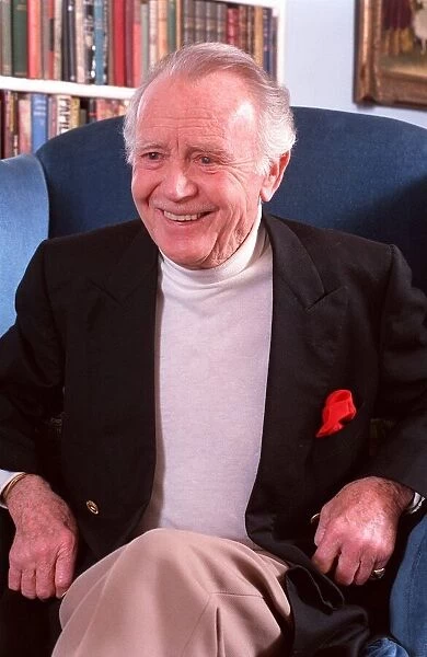 SIR JOHN MILLS IN PHOTO SHOOT AT HIS HOME SITTING IN ARMCHAIR WITH BOOKCASE AS BACKGROUND