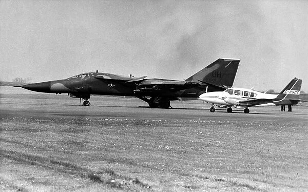 A USAF General Dynamics F-111 Aardvark sits on an airfield next to twin engined civil