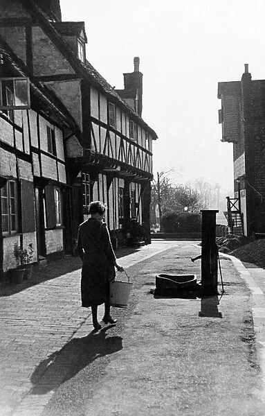 A woman carrying a bucket up the street to get water from a pump. February 1935