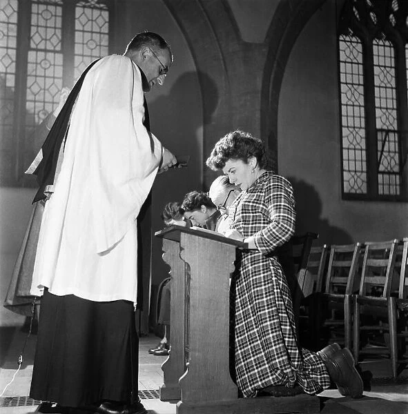 Woman kneeling at the altar praying in church watched by her priest December 1952