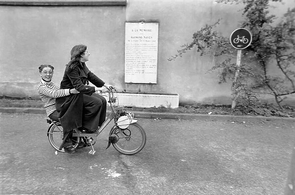Two women riding on an electric bicycle. April 1975 75-2072-021