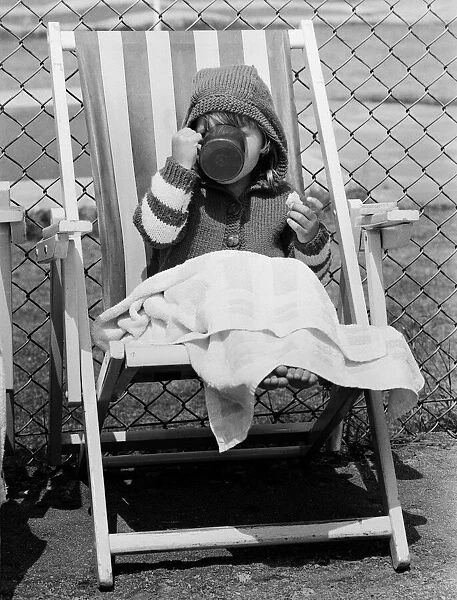 Young child swigs from a mug of juice, whilst sitting on a deck chair
