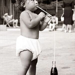 Baby drinking coke with a straw on the beach. Circa 1950