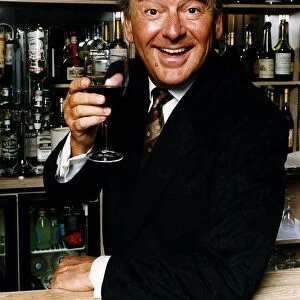 Bob Monkhouse raises a glass of wine standing behind a well stocked bar