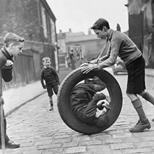 The boys are playing with a car tire, one boy is sitting inside the tire while another