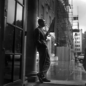 CARY GRANT SHELTERS FROM THE RAIN ON THE PORCH OF A LONDON HOTEL. 1957