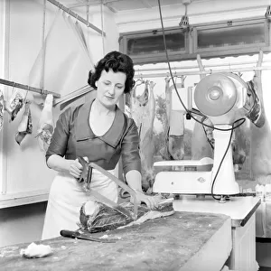 A female butcher seen here at work in her shop, cutting up a joint of meat. 1963