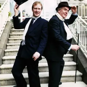 George Cole actor and Dennis Waterman standing on doorsteps raising a top hat and cane George Cole actor and Dennis Waterman standing on doorsteps raising a top hat and cane