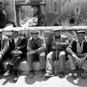 A group of pensioners sitting down against a wall on a street in a French village