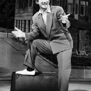 Johnny Beattie comedian comic actor on stage foot on suit case pointing back drop