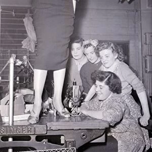 A lady in a pencil skirt stands on a bench while a lady sews on the sewing machine