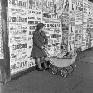 Little girl with toy pram stands waiting for her mother against a background of adverts Little girl with toy pram stands waiting for her mother against a background of adverts