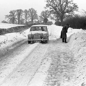A mini car drives along a snowy lane in the countryside near Coventry whilst a man tries