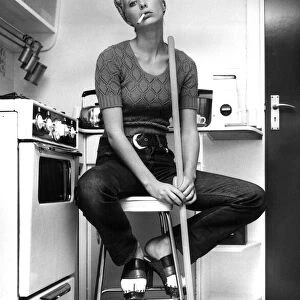 Model Nicki Howarth poses in the kitchen, holding a broom and smoking a cigarette