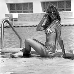 Model wearing a bikini and sunglasses as she sits by the side of a swimming pool in