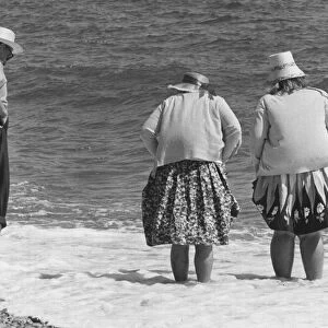 Two old women and one old man with hats stand on the beach with their feet in the water