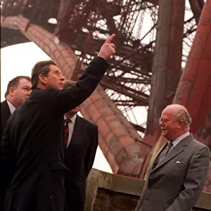 Prince Charles wearing black coat pointing finger to rust on Forth Rail Bridge