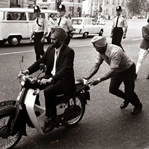 A Sikh man riding a motorcycle, wearing a turban. pushes by a fellow sikh