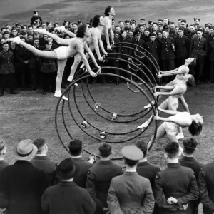 Women performing a gymnastic routine with large metal hoops, watched by military men Women performing a gymnastic routine with large metal hoops, watched by military men