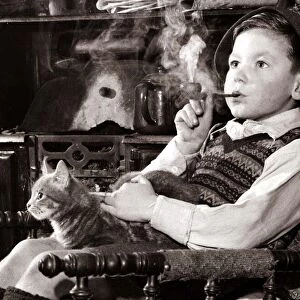 A young boy smoking a pipe and stroking a cat, sitting in a chair looking into