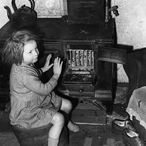 A young girl warms her hands by the oven. February 1957