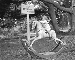Crying Prints: A baby riding a rocking horse beside a sign which reads No Horses by order. Circa 1940