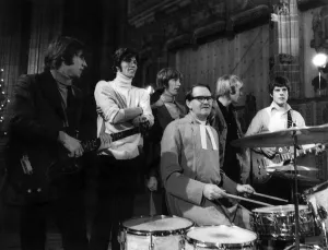The Bee Gees pop group stand behind a vicar having a a go on the drums during rehearsals