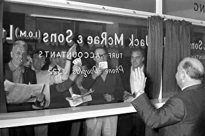 Bookmaker Sidney Glanz waves to punters waving cash at him through a glass window inside