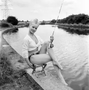 Clothing: Fashion: Beachwear: Model by the river bank fishing. August 1963 E490-005
